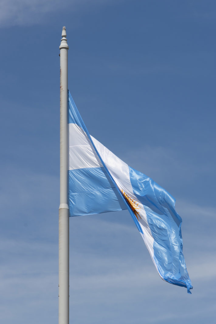 Argentine flag flying in Plaza de Mayo against a blue sky.