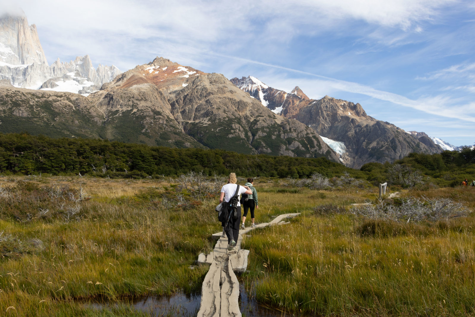 Walkers crossing narrow wooden boardwalks over marshy wetland on the Fitz Roy trail, with the mountains above El Chaltén in the background.