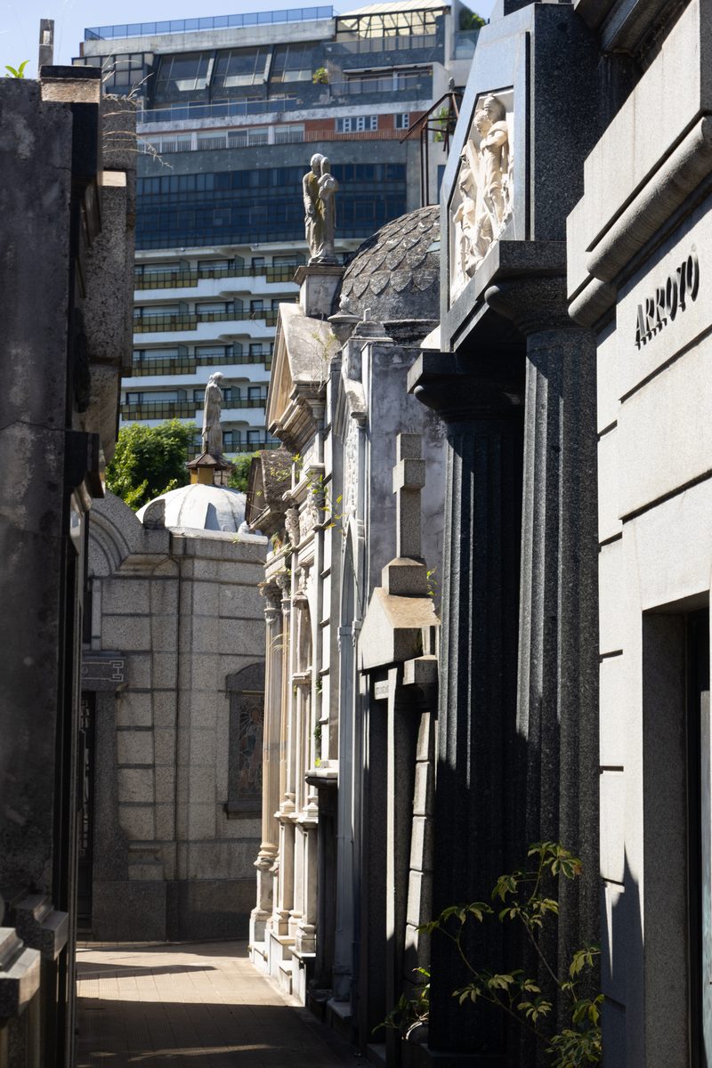 A narrow walkway lined with ornate mausoleums inside Recoleta Cemetery in Buenos Aires, with modern city buildings beyond.