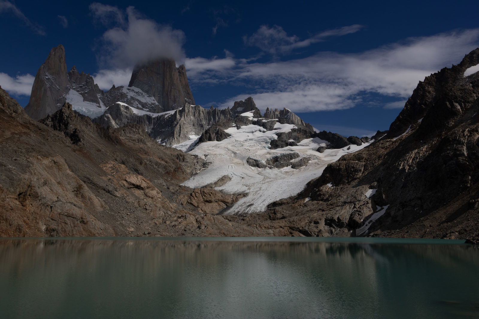 Laguna de los Tres below the granite towers and glaciers of Fitz Roy, with dark mountain slopes framing the turquoise lake in Patagonia.