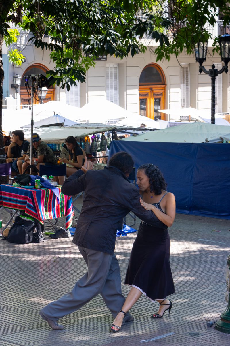 Couple dancing the tango in Plaza Dorrego, San Telmo, Buenos Aires, surrounded by market stalls and outdoor cafés.